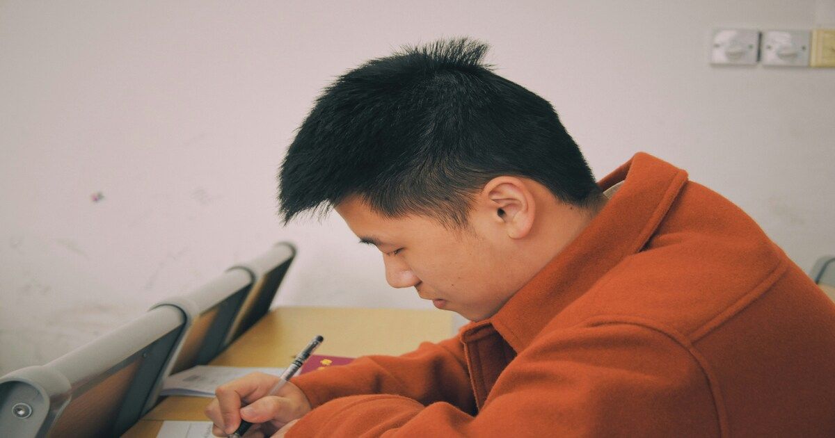 A teenage student sitting at a desk, writing in a notebook with a focused yet pensive expression