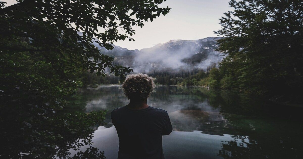 A man standing thoughtfully by the edge of a calm river, surrounded by natural scenery.