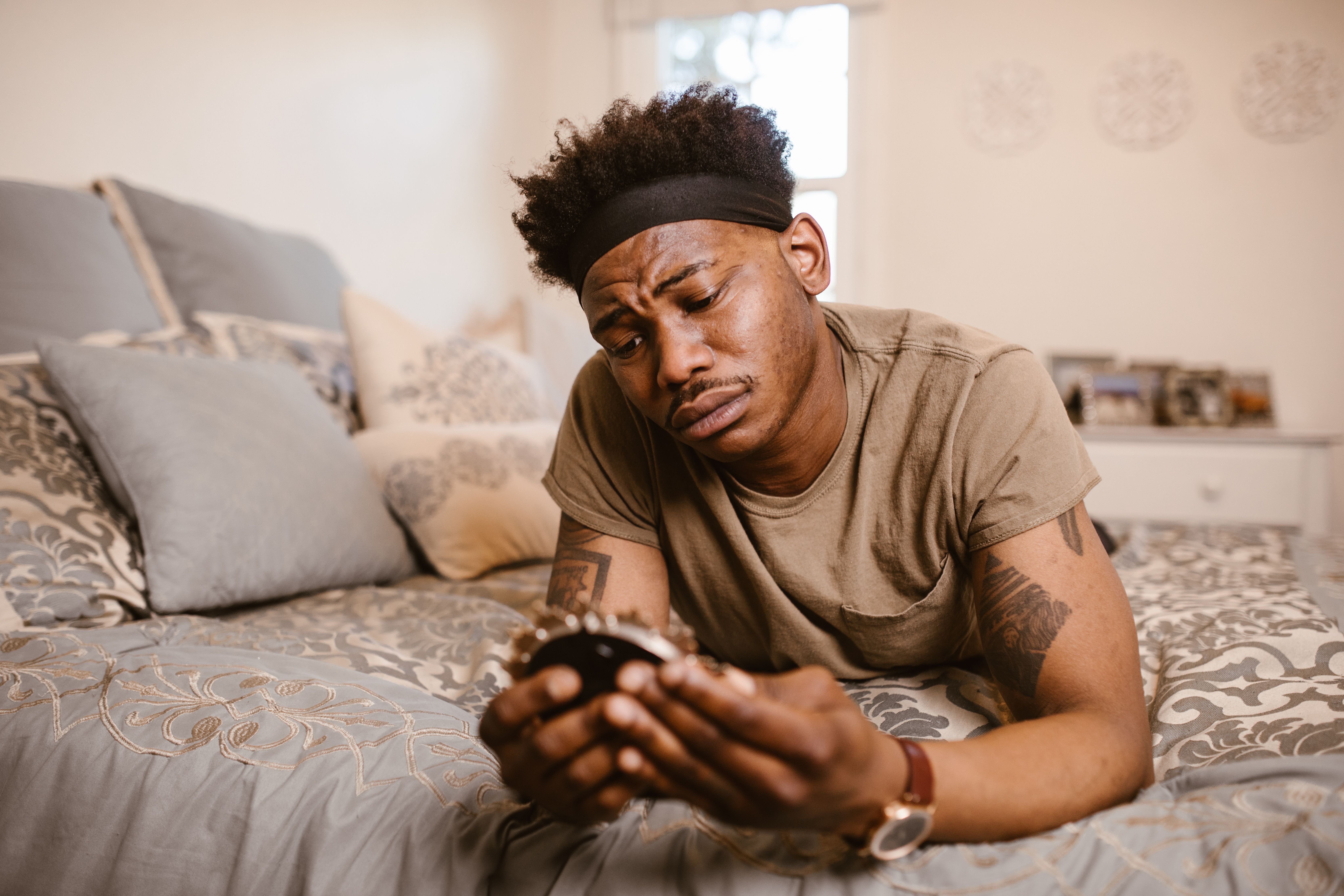 Man lying on a bed in a tan t-shirt and black headband, looking worried at a small round clock held in both hands.