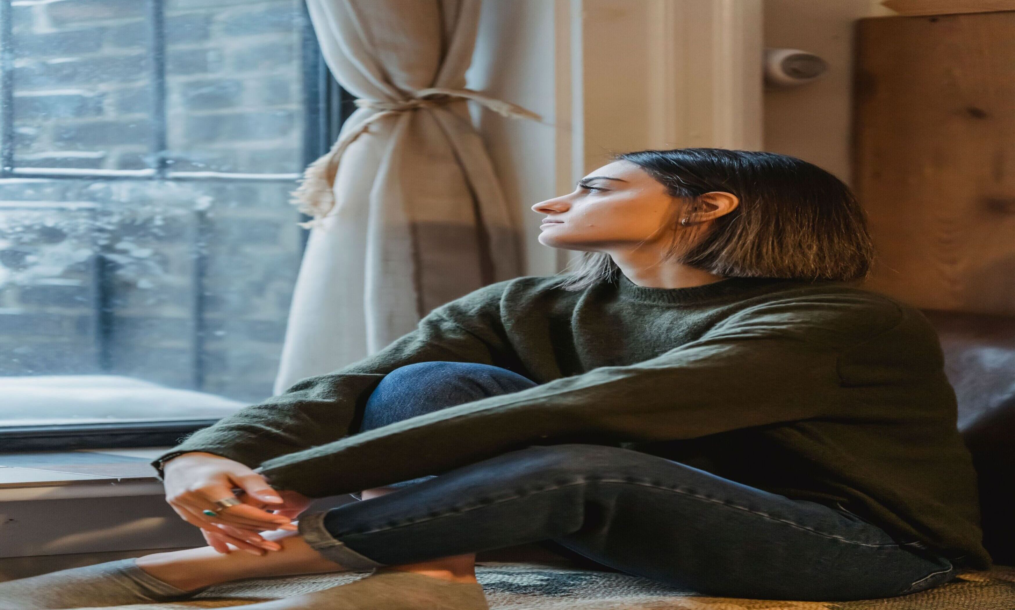 Woman in a green sweater sitting on the floor by a window with her arms around one knee, looking outside.