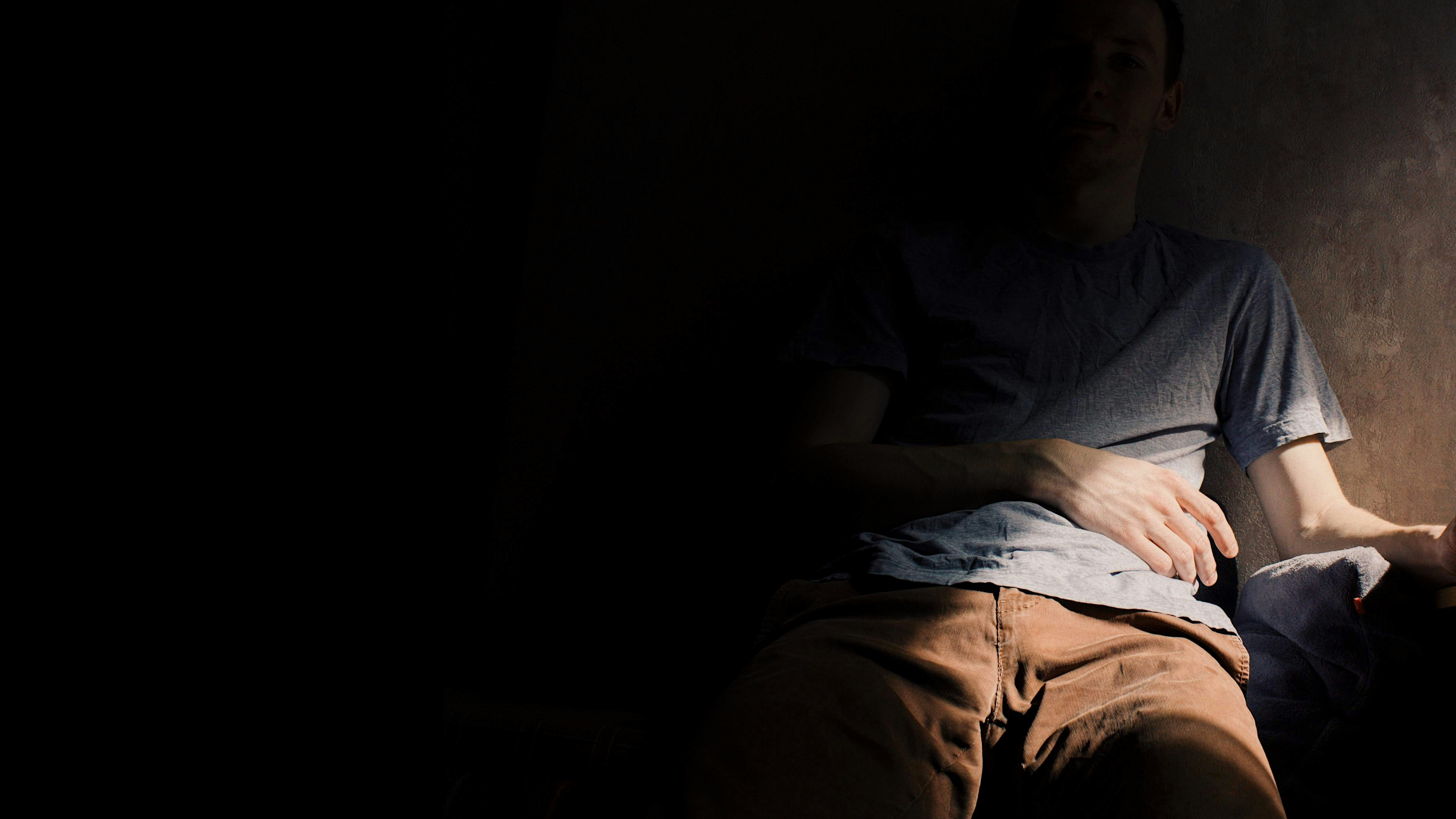 A person sitting in a dark room with a hand resting on their stomach.